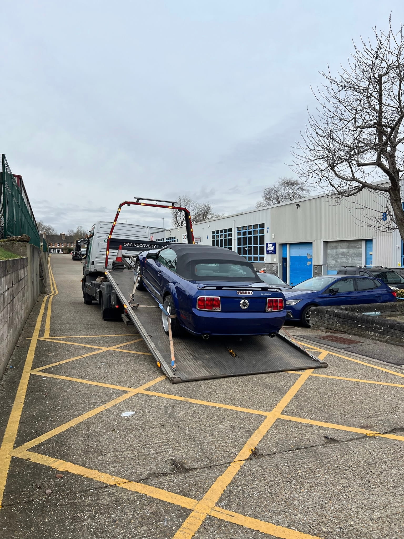 A blue convertible car is being loaded onto a flatbed tow truck labeled "CAR RECOVERY" outside a large garage, against an overcast sky.