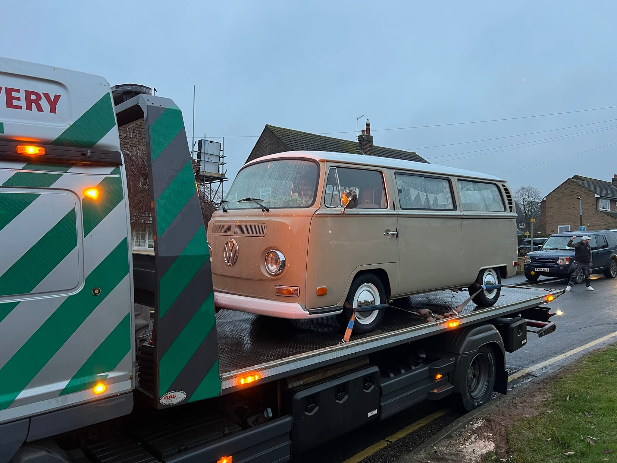 A beige vintage Volkswagen van is loaded onto a recovery truck on a wet street. Overcast sky, streetlights glow, suggesting a calm, rainy evening.