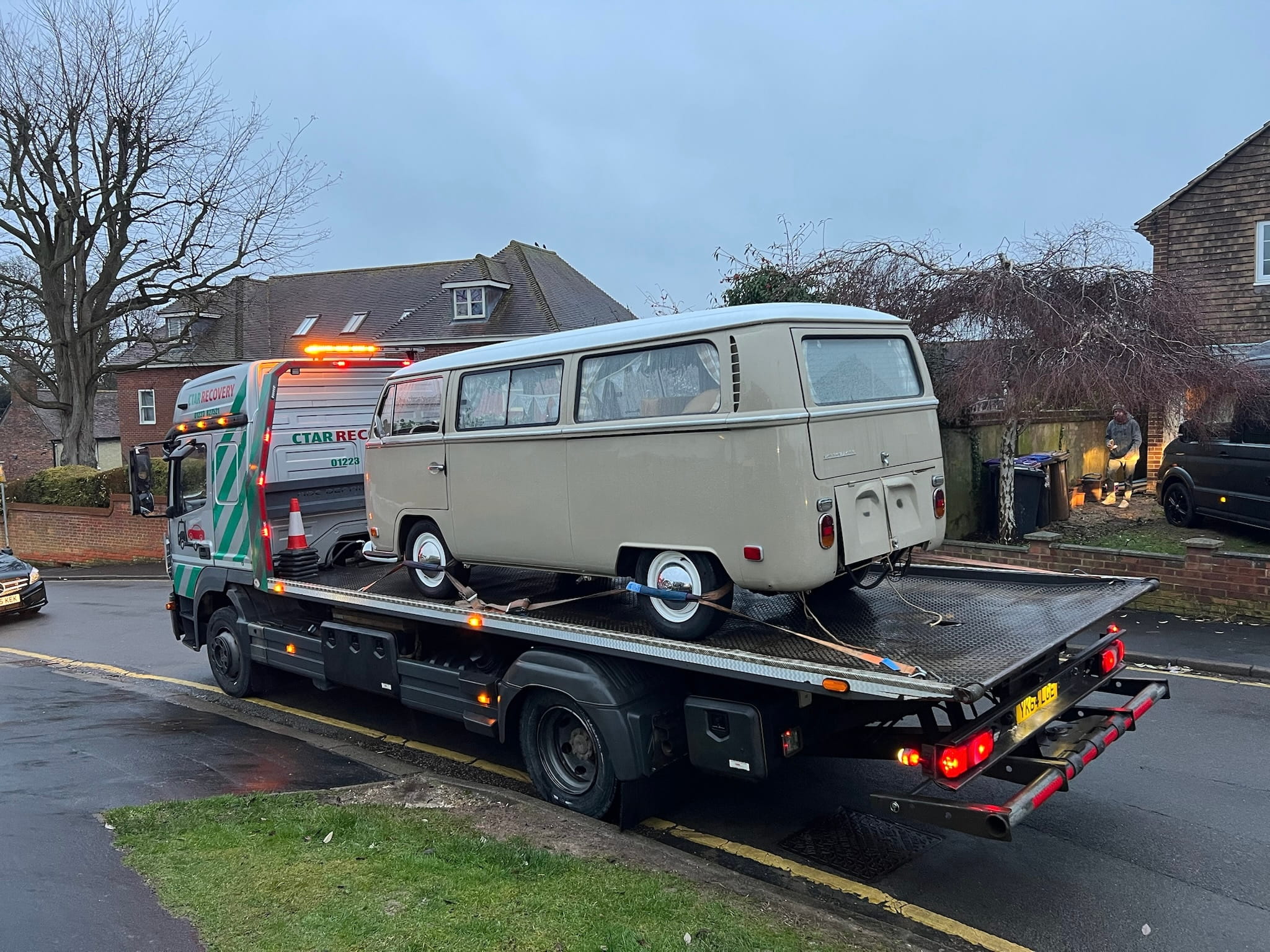 A vintage beige van is secured on a flatbed tow truck on a residential street. It's overcast and the surrounding houses have a cozy, suburban feel.