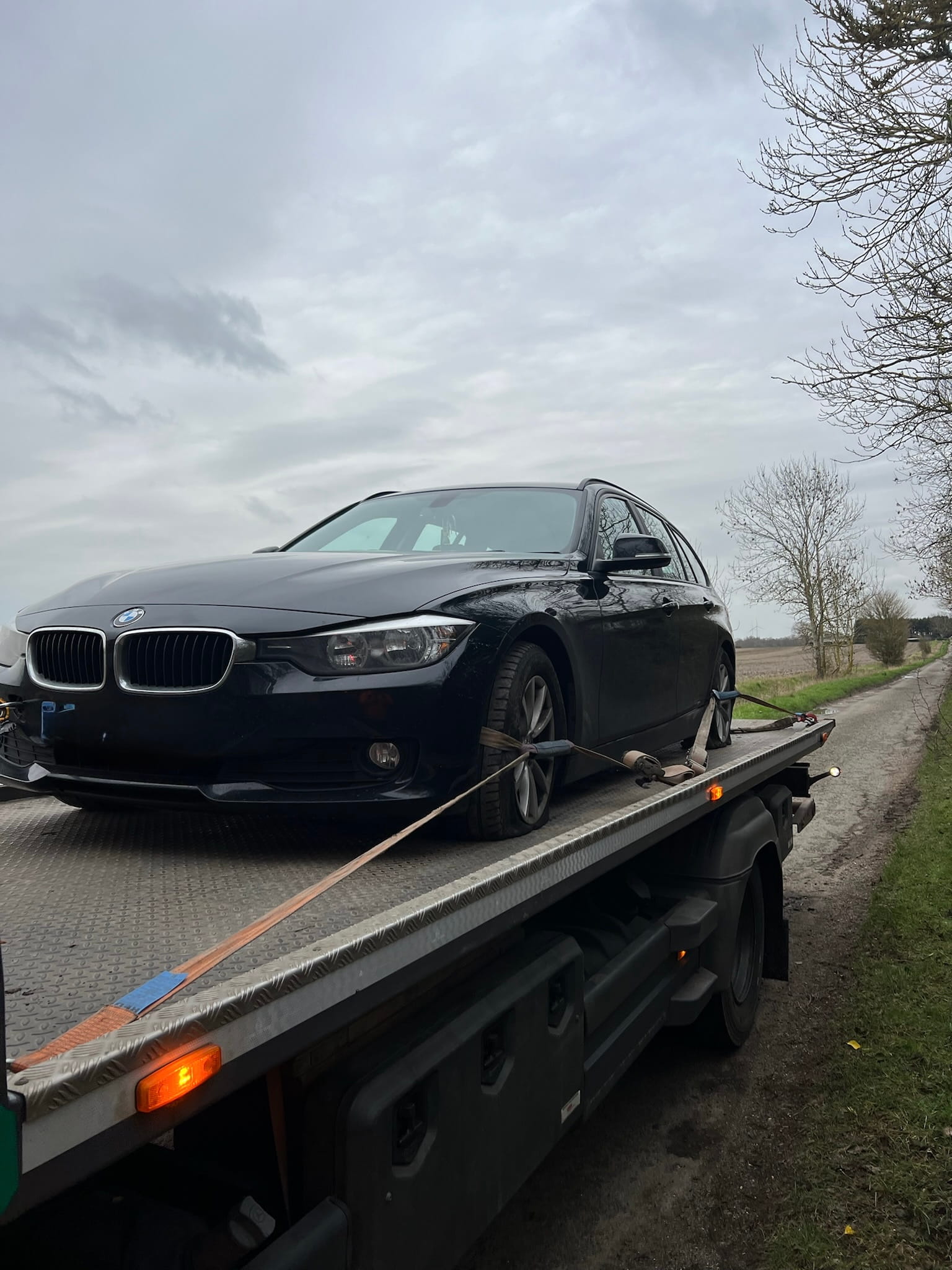 A black car secured on a flatbed tow truck on a rural road. Overcast sky and bare trees in the background convey a calm, dreary atmosphere.